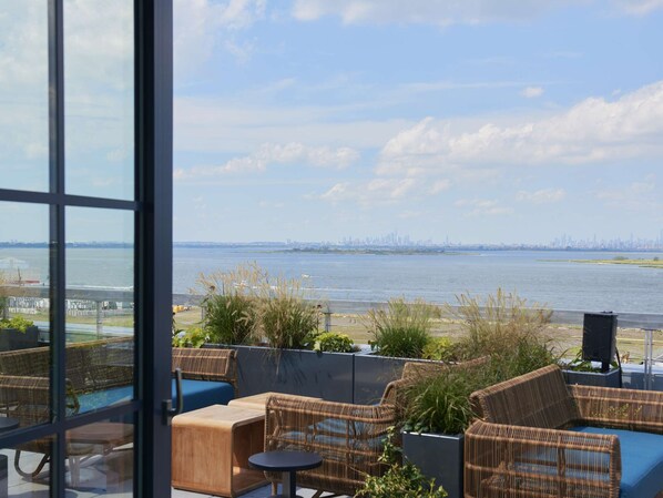 A terrace with wicker furniture and a view of a city skyline across a body of water. - Rockaway Beach Stay | Pool. Rooftop. Dining + Spa (Queens)