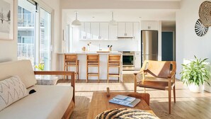 A modern kitchen with white cabinets, a central island with bar stools, a dining table with books, and a sofa. - Rockaway Beach Stay | Pool. Rooftop. Dining + Spa (Queens)