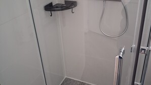 Een douchecabine met glazen wanden, een handdouche, een zwart plankje en een betegelde vloer.