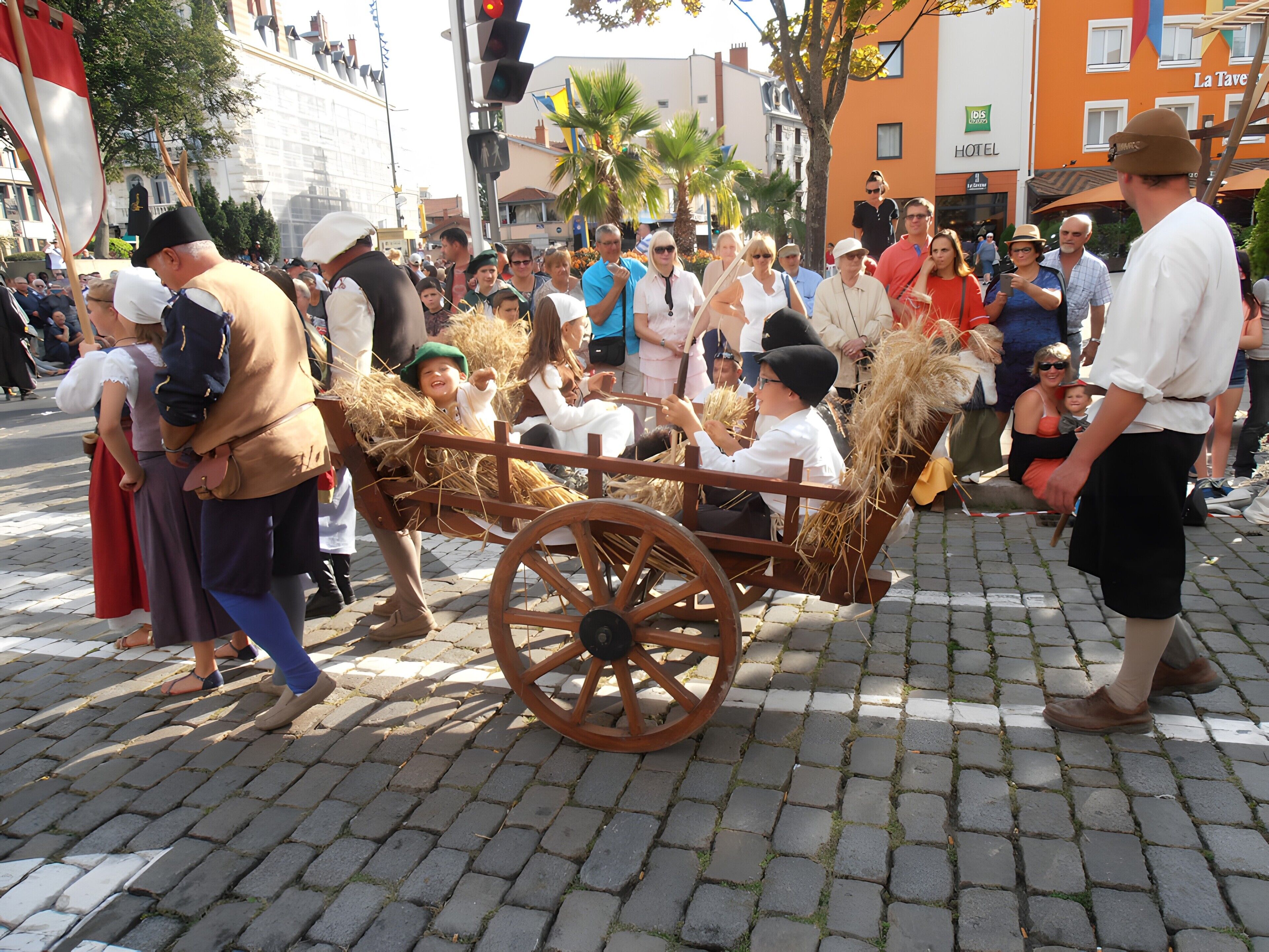 A cobblestone street with a crowd of people, some in traditional attire, and a wooden cart with hay.