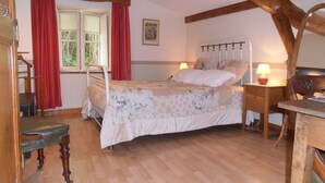 A bedroom with a wooden bed frame, a bedside table with a lamp, a chair, and a window with curtains. - Guest Room “Chambre Rêverie” with Shared Terrace, Garden and Wi-Fi (Polignac)