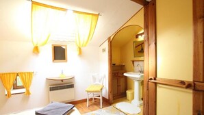 A room with a bed, a chair, a small table, a window with curtains, a sink, and a mirror.