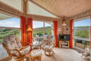 A wooden interior with wicker furniture, a television, and large windows overlooking a grassy landscape. - Lovely holiday home with covered terrace, only 300 meters from the North Sea in. (Ringkøbing)