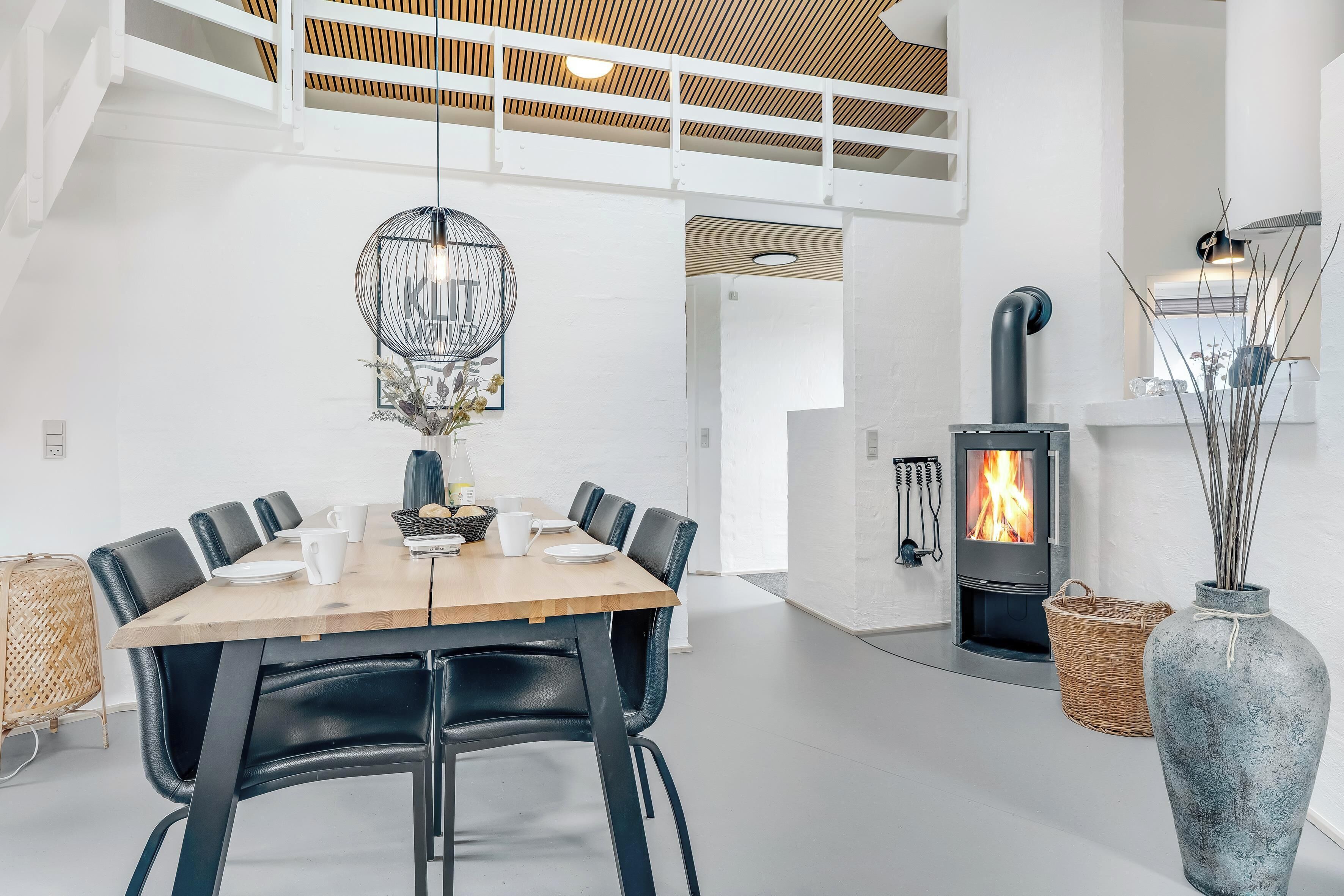 Un interior moderno con una mesa de comedor, sillas, una lámpara colgante, una chimenea y un jarrón grande.