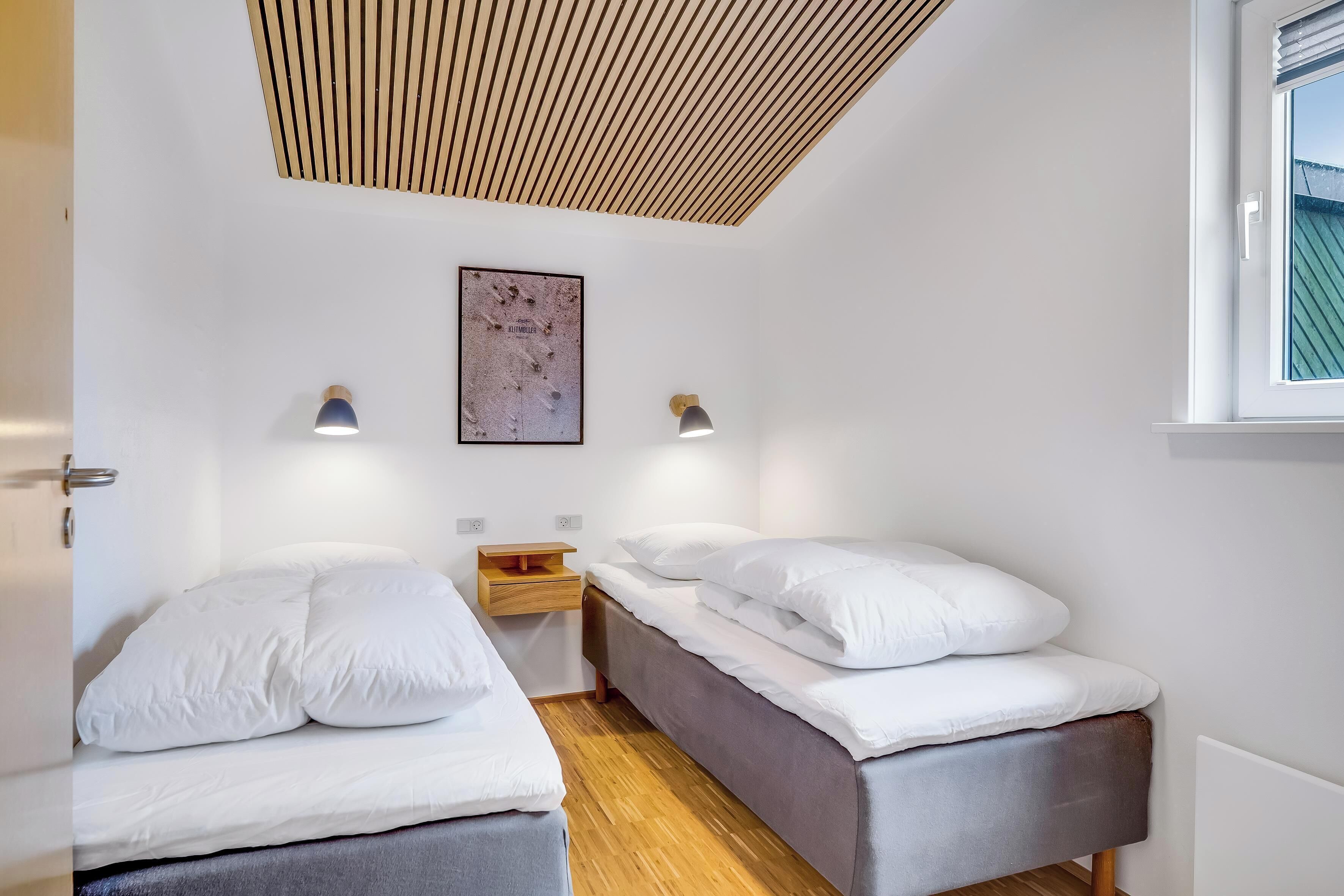 Quarto com duas camas, teto de ripas de madeira, luminárias fixadas na parede e janela com persianas.