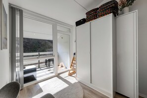 Una habitación con puerta de vidrio corrediza que da a un balcón, un armario blanco y una pequeña silla de madera.