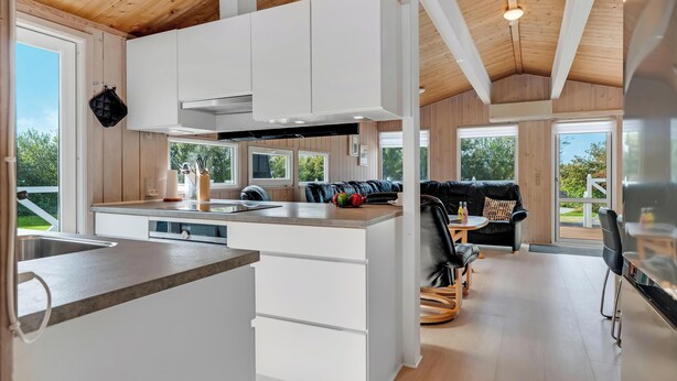 A modern kitchen with white cabinetry, a central island, and a dining area with a black leather couch and wooden table. - Lovely holiday home with annex and wilderness bath. (Vestervig)