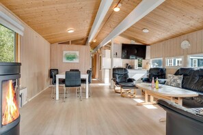 Un intérieur moderne avec un plafond en bois, un coin repas comprenant des chaises et une table, un canapé en cuir noir, une cheminée et un coin cuisine avec un téléviseur.