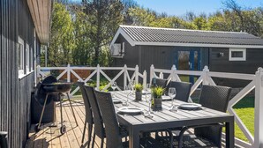 An outdoor deck with a table set for dining, surrounded by chairs, and a grill. - Lovely holiday home with annex and wilderness bath. (Vestervig)