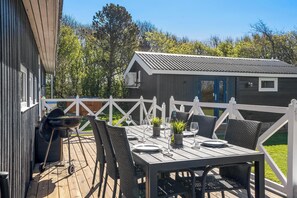 An outdoor deck with a table set for dining, surrounded by chairs, and a grill. - Lovely holiday home with annex and wilderness bath. (Vestervig)