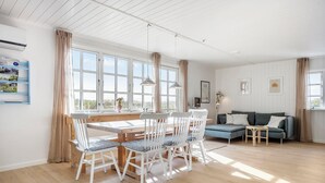 Ruime kamer met een houten eettafel, witte stoelen, een blauwe bank en een groot raam met gordijnen.