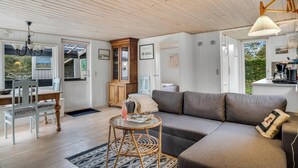 Ein gemütliches Wohnzimmer mit einer Couch, einem Esstisch mit Stühlen, einem Korbstuhl-Beistelltisch und einer durchgehenden Tür erkennbaren Küchenzeile.