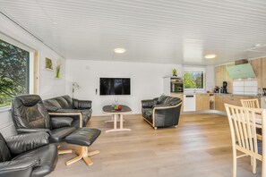 A modern living room with leather sofas, a wooden dining table, and a kitchen area with wooden cabinets and a built-in oven.
