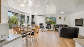 Een ruime woonkamer met een eettafel, stoelen, een bank en een aan de muur bevestigde televisie.