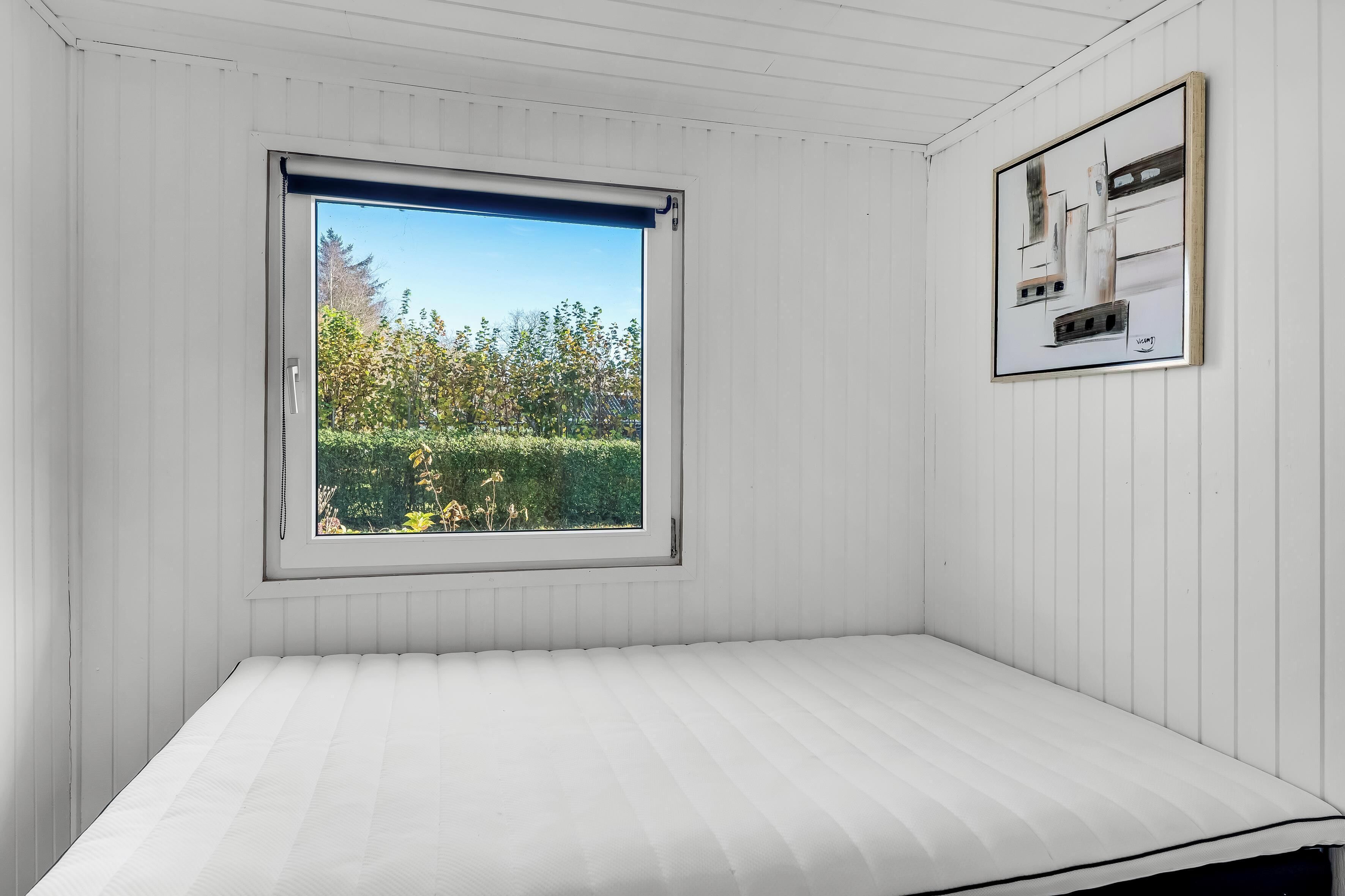 A room with a bed, a window with a view of greenery, and a framed picture on the wall.