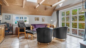 Een kamer met een houten plafond, een groot raam met uitzicht op bomen, een paarse bank en een zwarte lederen fauteuil.