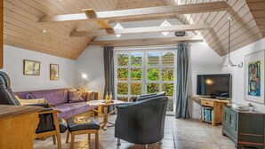 Een gezellige woonkamer met een houten plafond, een bank, een zwarte fauteuil, een televisie en een raam met gordijnen.