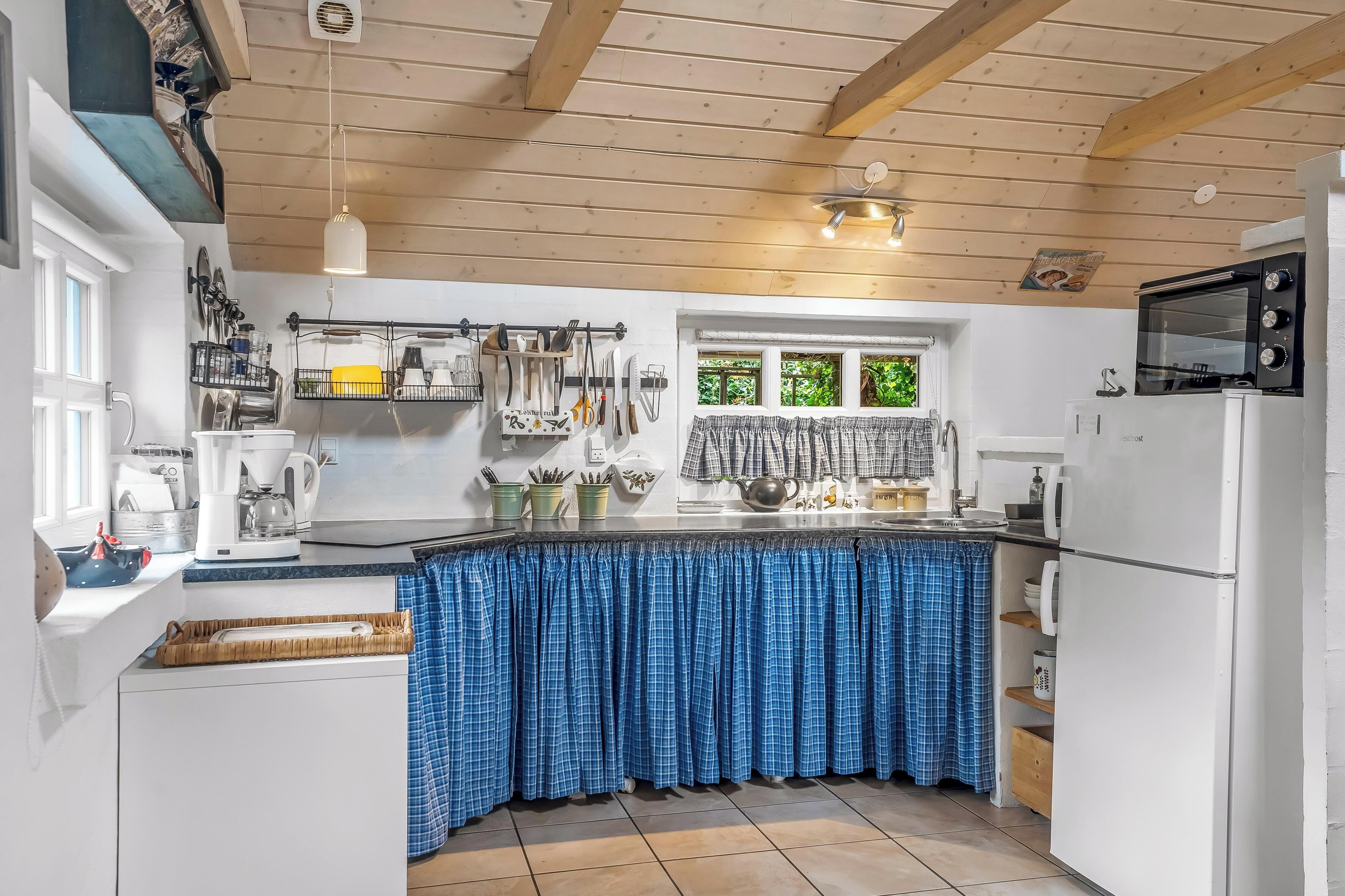 A kitchen with a wooden ceiling, white cabinets, a blue checkered tablecloth, and a window with greenery outside.