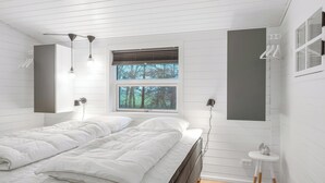 Ein Schlafzimmer mit Bett, zwei Nachttischlampen, einem Fenster mit Blick auf Bäume, einem kleinen weißen Tisch und einem Wandregal.