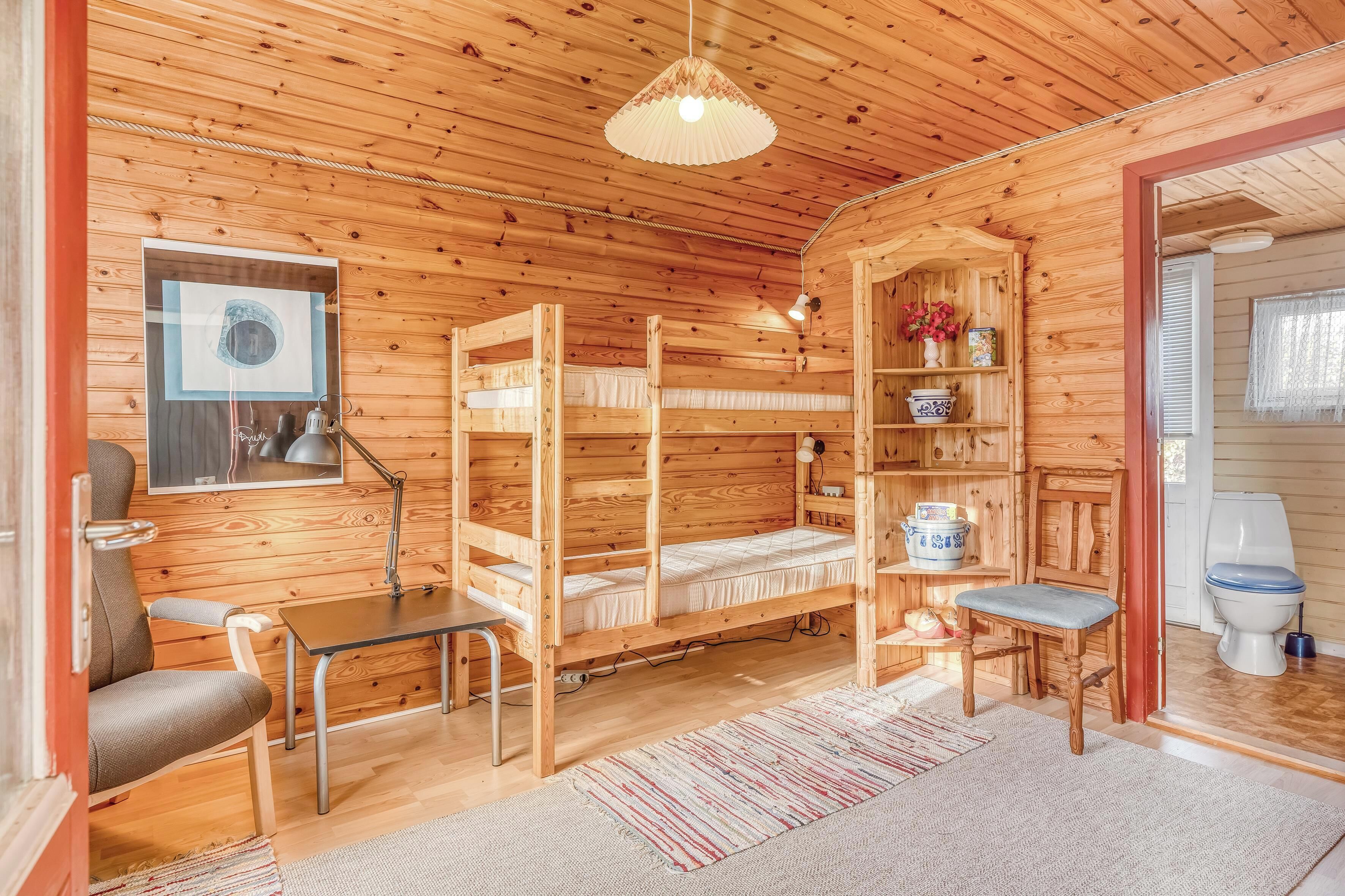 A wooden cabin interior with bunk beds, a desk, a chair, a lamp, a shelf with decorative items, and a toilet.