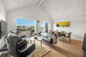 A spacious living area with a modern sofa, a dining table, and a large window offering a view of the outside. - Lovely holiday apartment from 2026 in Søndervig, close to the North Sea. (Ringkøbing)