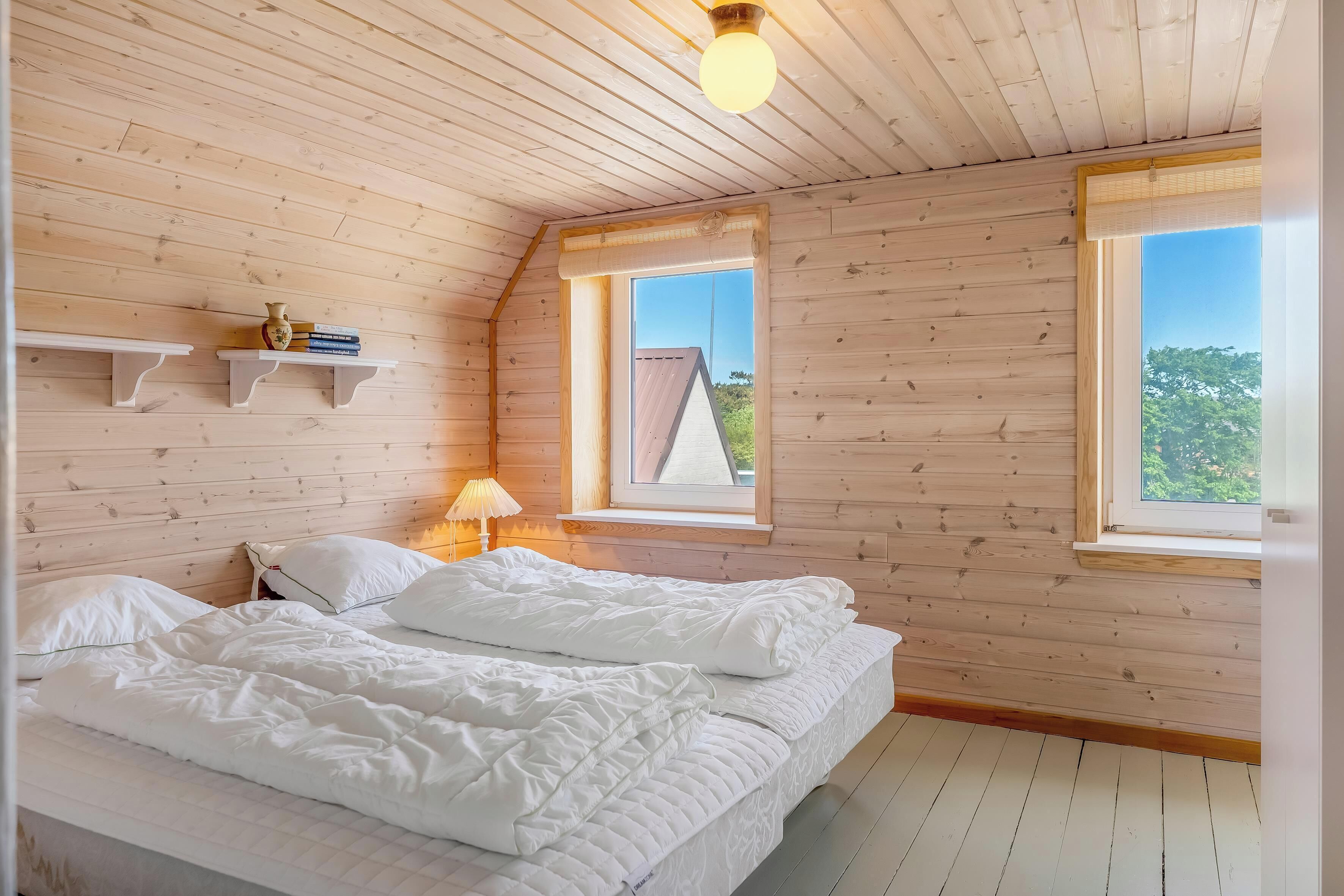 A bedroom with a wooden ceiling, a bed with white bedding, two windows, and a nightstand with a lamp.