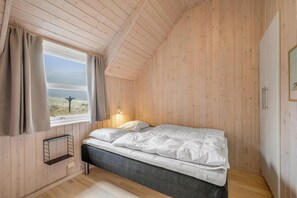 Ein Schlafzimmer mit Holzboden, einem Bett mit weißer Bettwäsche, einem Fenster mit Vorhängen und einem Nachttisch.