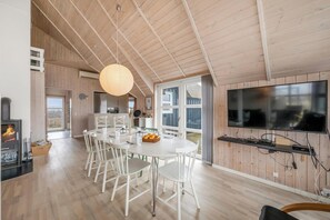 Ein modernes Interieur mit Holzboden, einem Essbereich mit weißen Stühlen, einem an einer Holzwand montierten Fernseher und einem Kamin.