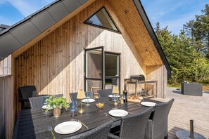 An outdoor dining area with a wooden deck, a table set for two, and a grill in the background. - Lovely holiday home built in 2025, with space for 8 people close to the North. (Ringkøbing)