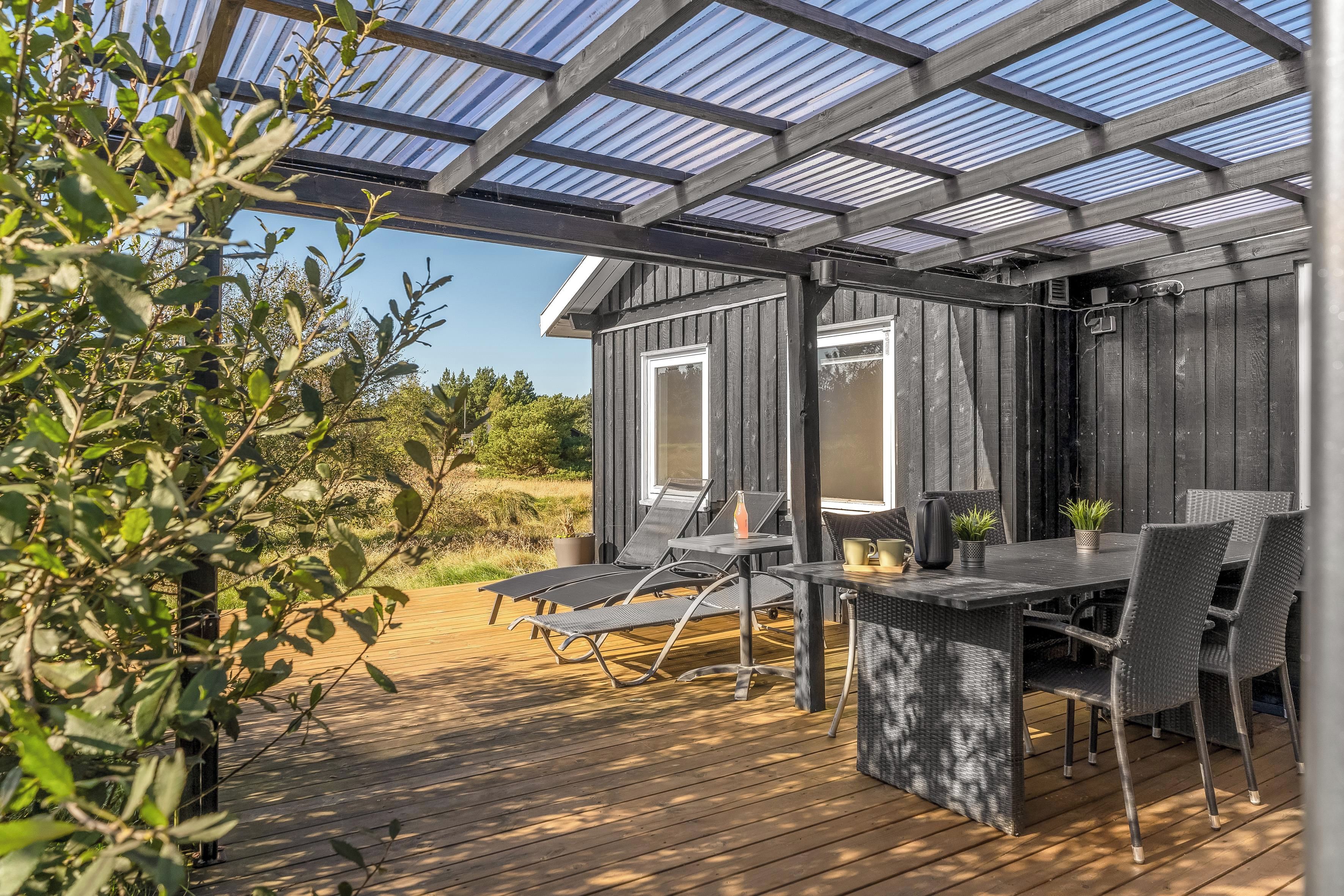 An outdoor patio with a wooden deck, metal chairs, a table, and a glass-covered pergola structure.