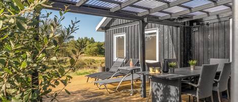An outdoor patio with a wooden deck, metal chairs, a table, and a glass-covered pergola structure.