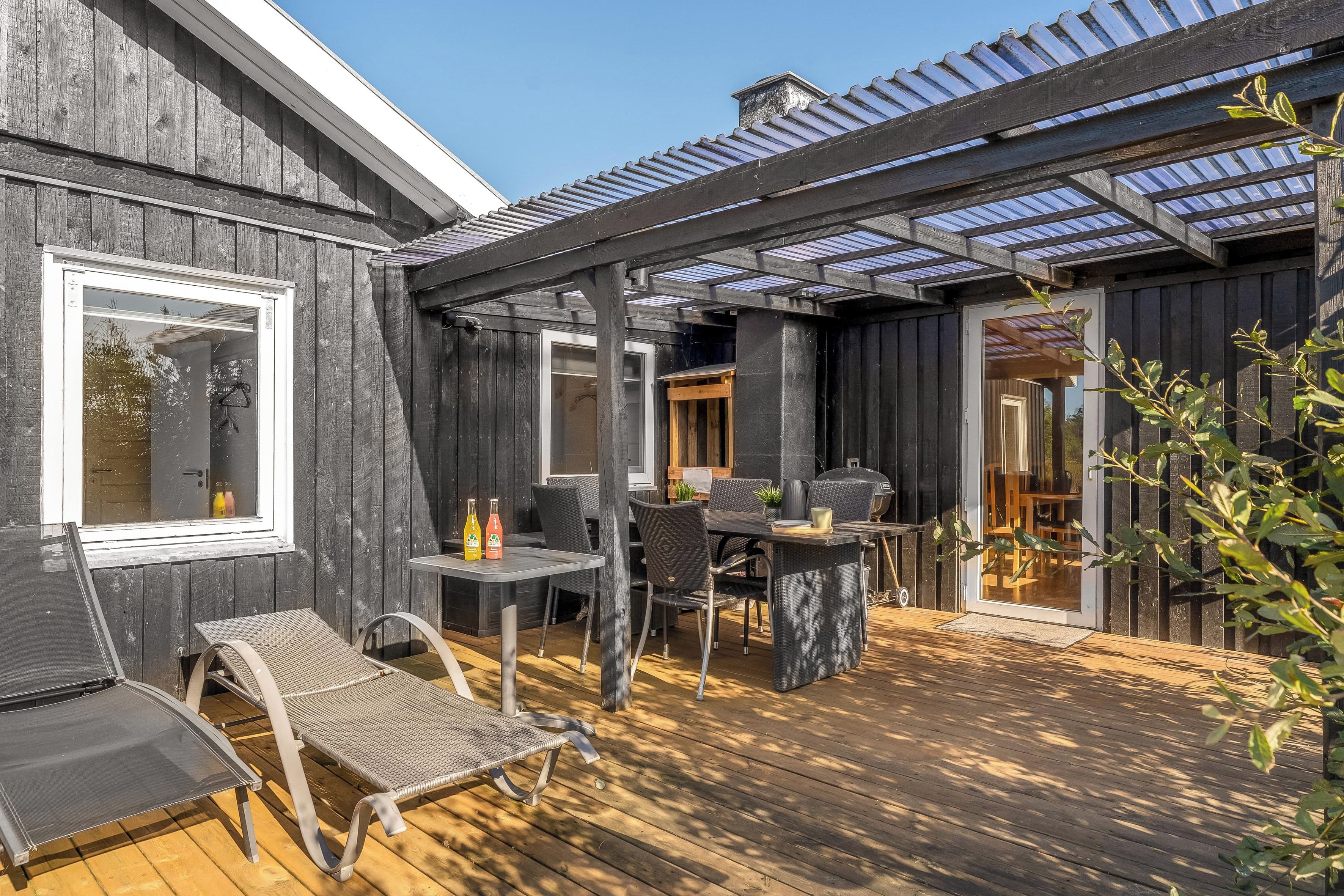 A wooden deck with outdoor furniture, a pergola with glass panels, and a view of the interior.