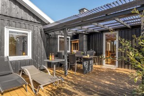 Una terraza de madera con mobiliario exterior, una pérgola con paneles de vidrio y vista al interior.