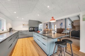 A modern kitchen with a large island, bar stools, and a dining area with a brick wall and wooden furniture. - Modernized holiday home with enclosed terrace, near Stauning harbor (Skjern)