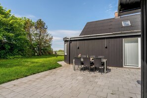 Een terras met een tafel en stoelen, een gazon en een gebouw met een donkere gevel en een raam.