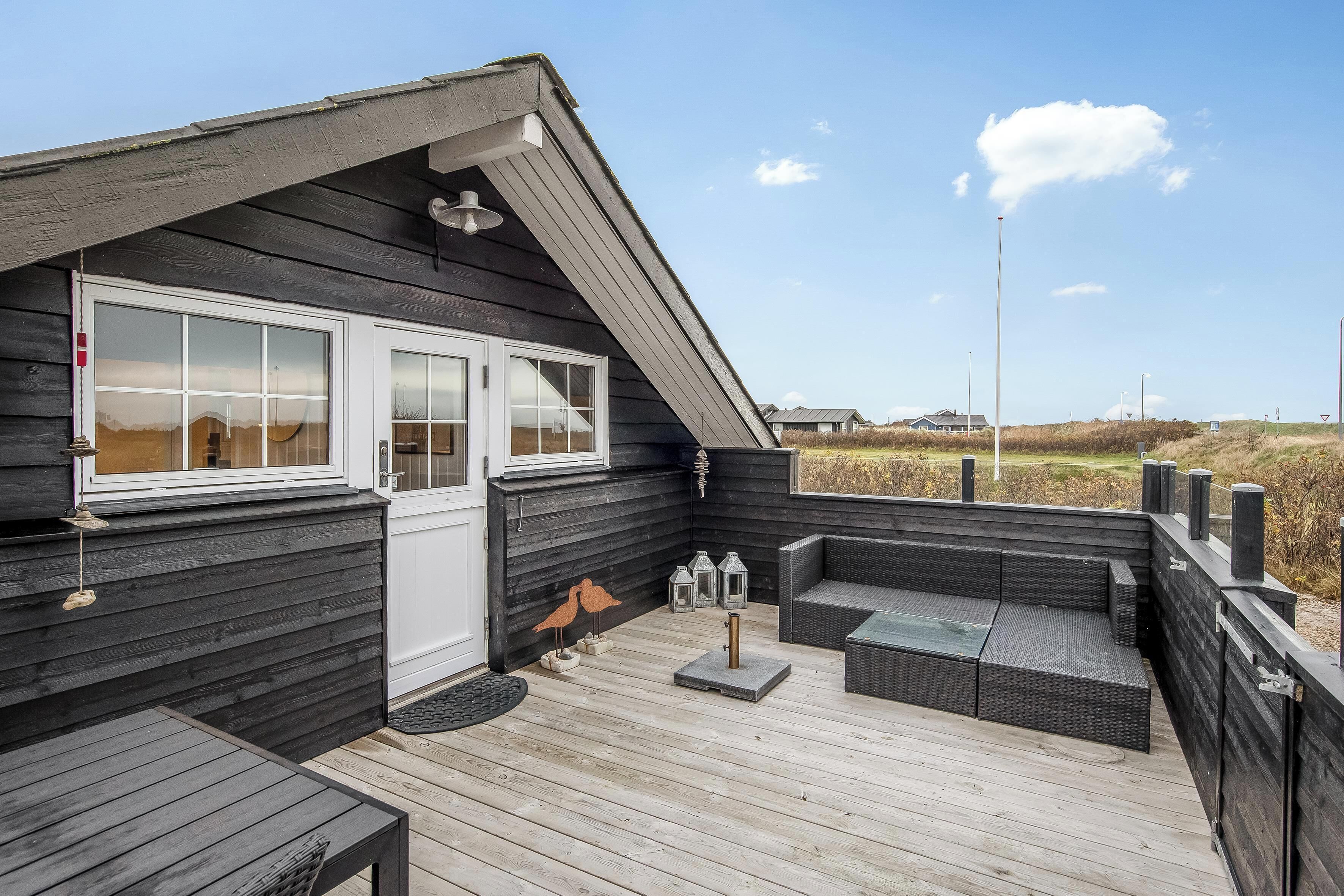 Una terraza de madera con mobiliario exterior, una puerta blanca y vistas al paisaje.