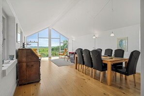Un vaste espace de salle à manger avec une table et des chaises en bois, de grandes fenêtres offrant une vue sur l’extérieur et un luminaire suspendu moderne.