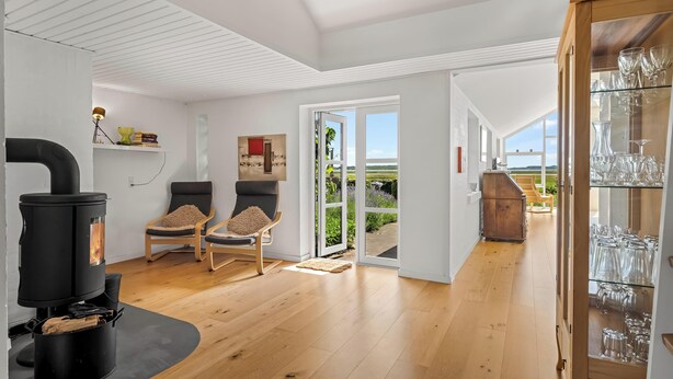 Un intérieur moderne avec un poêle à bois, deux fauteuils, une vitrine contenant de la verrerie, et une vue sur un jardin extérieur, visible à travers une porte vitrée.