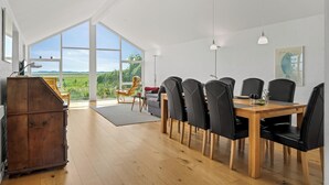 Un vaste espace de salle à manger avec une table en bois et des chaises noires, de grandes fenêtres offrant une vue sur la verdure, et un luminaire suspendu moderne.