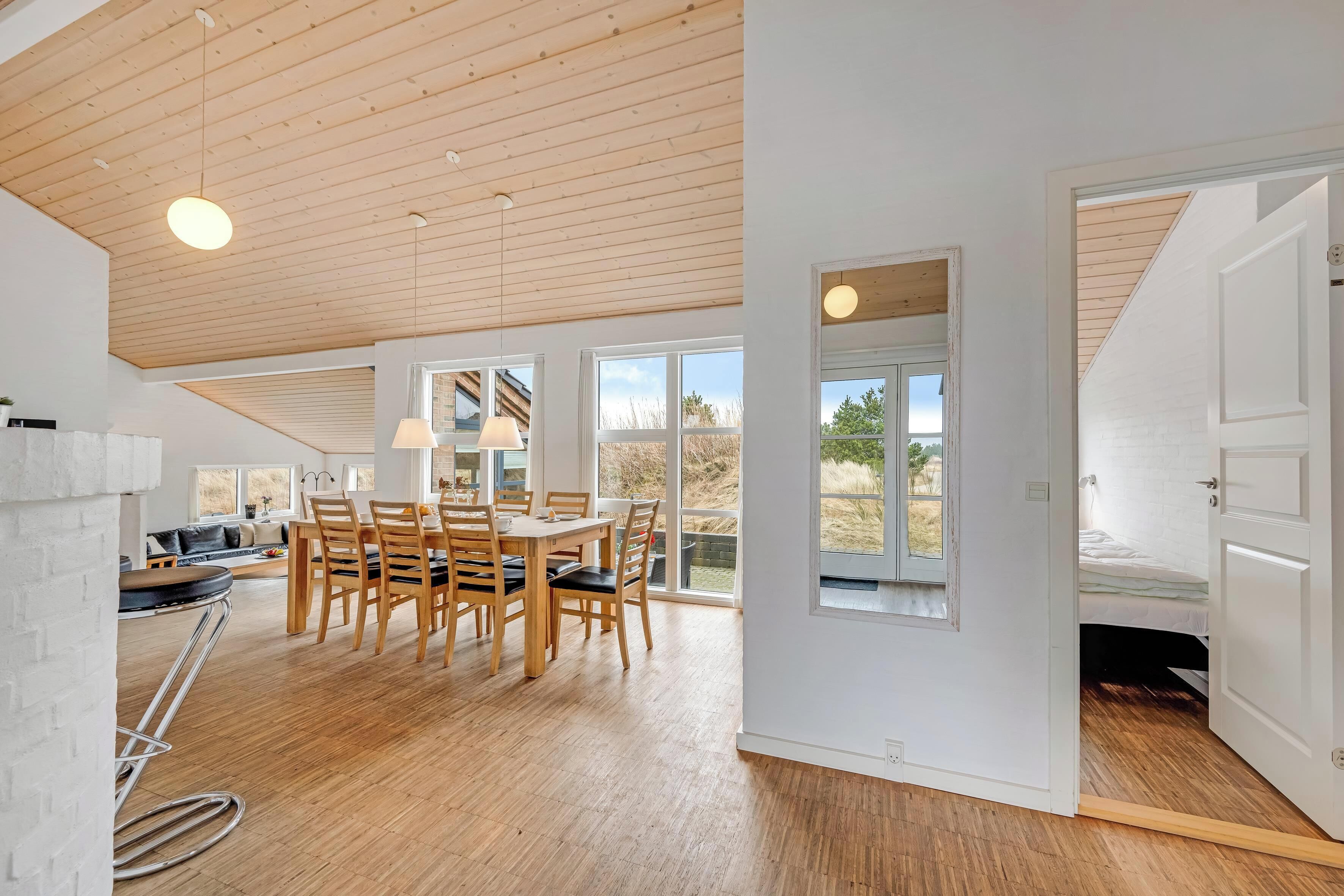 Een ruime kamer met een hoge plafond, houten vloer, een zithoek met stoelen en een tafel, en grote ramen met uitzicht op het omliggende landschap.