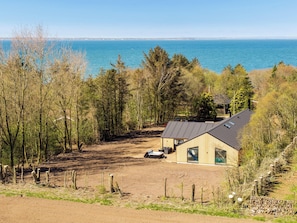 A house with a metal roof, surrounded by trees and a dirt path, overlooking a body of water.