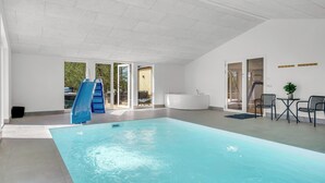 An indoor swimming pool with a slide, a seating area with chairs, and a view of the outdoors through glass doors.