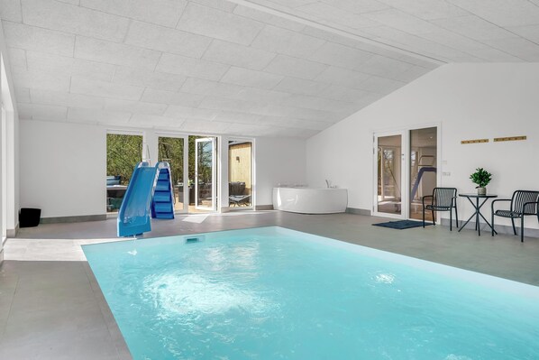 An indoor swimming pool with a slide, a seating area with chairs, and a view of the outdoors through glass doors. - Luxury pool house for 20 people with activity room and 6-person outdoor spa (Spøttrup)