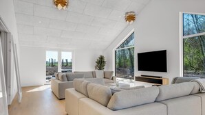 A spacious living room with a large sectional sofa, a flat-screen TV, and a large window offering a view of trees.