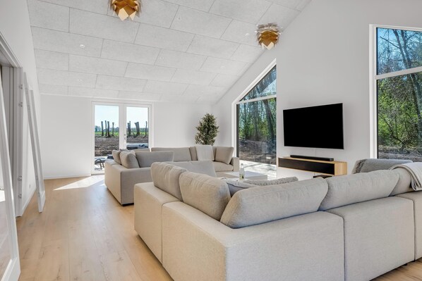 A spacious living room with a large sectional sofa, a flat-screen TV, and a large window offering a view of trees. - Luxury pool house for 20 people with activity room and 6-person outdoor spa (Spøttrup)