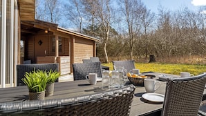 Een terras met een eethoek, rieten stoelen, een tafel gedekt met borden, bekers en een mand met brood.