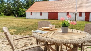 Une table ronde en osier, avec des chaises, un plateau contenant des assiettes et des tasses, et une plante en pot ornée de fleurs.