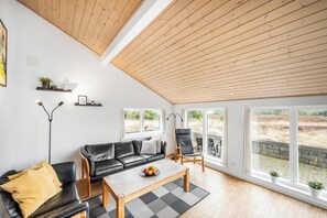 Un salon moderne avec un canapé en cuir noir, une table basse en bois et un tapis noir et blanc. On y trouve deux fauteuils noirs, un lampadaire et une fenêtre donnant sur l’extérieur.