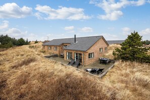 Une maison à deux étages, construite en briques et dotée d’un toit en tuiles, avec une terrasse meublée et un jardin verdoyant tout autour.