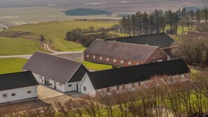 Eine ländliche Landschaft mit einem großen Bauernhaus und angrenzenden Gebäuden, umgeben von offenen Feldern und sanften Hügeln in der Ferne.